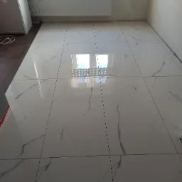 Błyszcząca podłoga z białych płytek imitujących marmur, świeżo położona w pustym pomieszczeniu z grzejnikiem pod oknem, widoczne fugi i odbicie okna w płytkach.