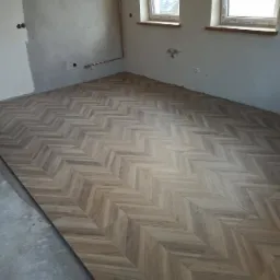 Ułożona pod kątem jodełki podłoga z paneli imitujących drewno w surowym wnętrzu z widocznymi instalacjami i oknami.