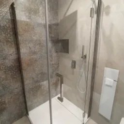 Kabina prysznicowa z dekoracyjnymi płytkami ściennymi w odcieniach szarości i brązu, szklane drzwi z chromowanym uchwytem, biały brodzik i grzejnik łazienkowy z ręcznikiem.