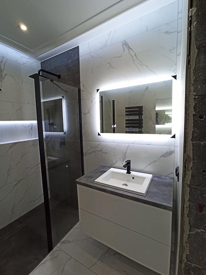 Wykończona łazienka z kabiną prysznicową typu walk-in, umywalką nablatową na szafce z szufladami, podświetlanym lustrem i płytkami imitującymi marmur na ścianach i podłodze, widoczne niedokończone...