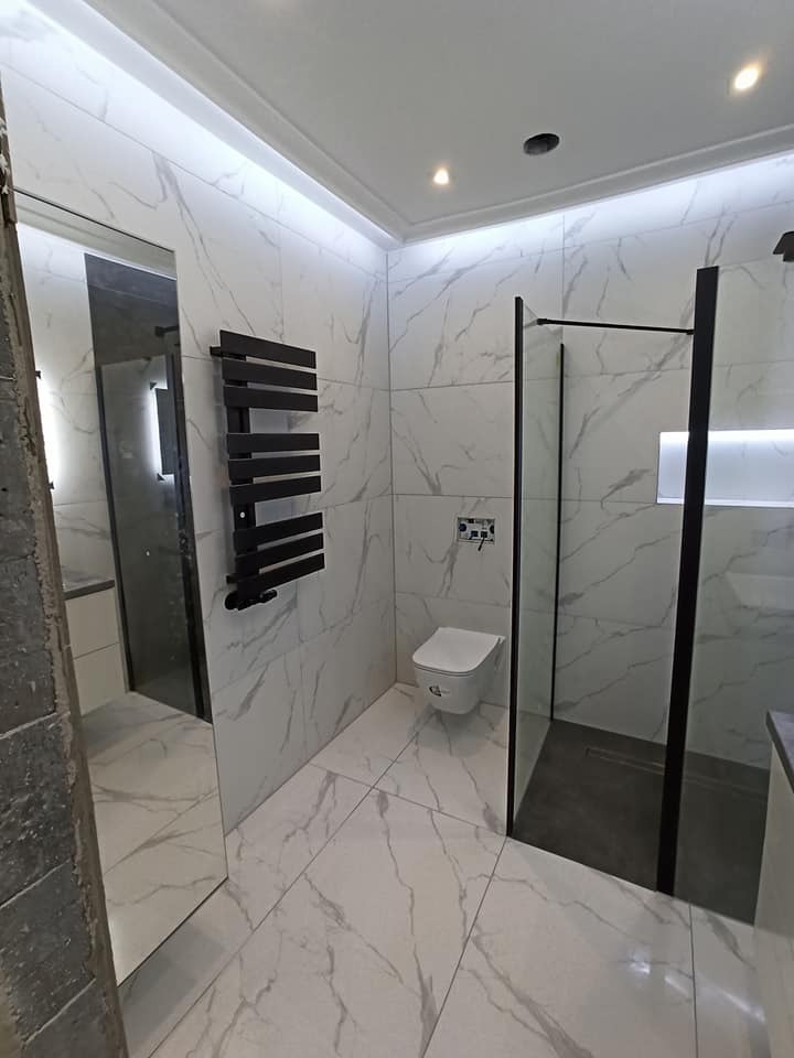 Wykończona łazienka z marmurowymi płytkami, czarnym grzejnikiem drabinkowym i kabiną prysznicową z czarnymi ramami, widok z narożnika pomieszczenia, częściowo zasłoniętego przez element konstrukcyjny.