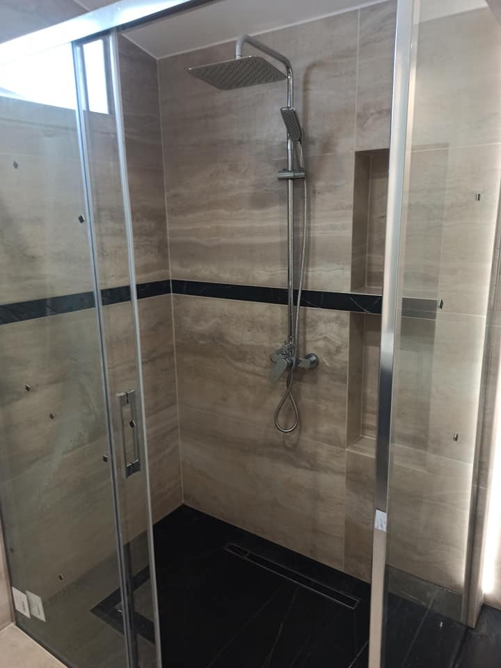 Kabina prysznicowa z deszczownicą i czarną listwą dekoracyjną na beżowych płytkach imitujących marmur, czarna podłoga z odpływem liniowym, półka w ścianie.