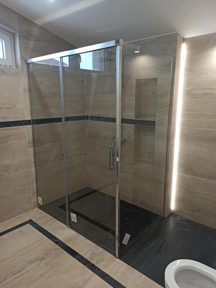 Szklana kabina prysznicowa z przesuwnymi drzwiami w łazience wykończonej płytkami imitującymi marmur, z czarnym pasem dekoracyjnym na ścianach i podłodze.