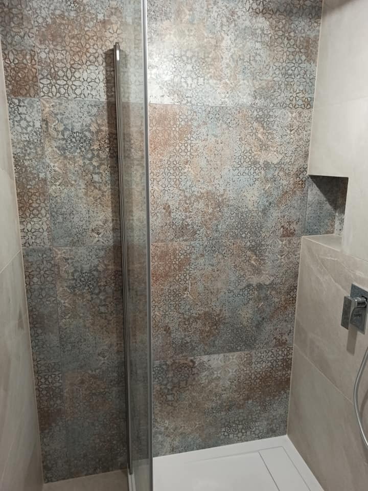 Kabina prysznicowa z ozdobnymi, wzorzystymi płytkami w odcieniach szarości, brązu i beżu, wbudowana półka w ścianie, bateria prysznicowa z termostatem.