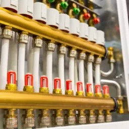 Rozdzielacz ogrzewania podłogowego z białymi rurami i czerwonymi przepływomierzami, zamontowany w szafce instalacyjnej.