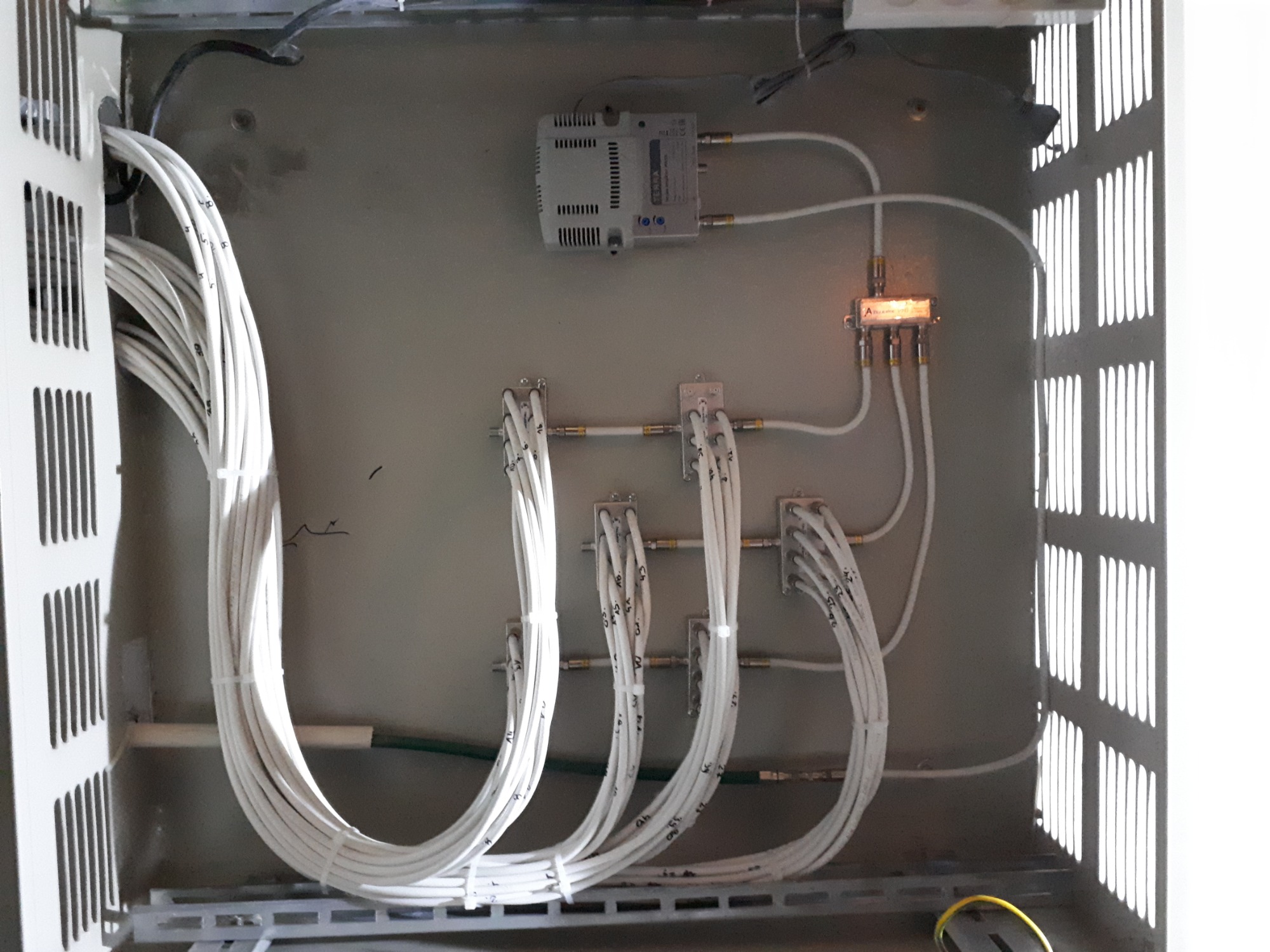 Wnętrze szafy telekomunikacyjnej z ułożonymi kablami koncentrycznymi, oznaczonymi numerami, podłączonymi do rozgałęźników i wzmacniacza sygnału. Widoczne metalowe elementy konstrukcyjne...