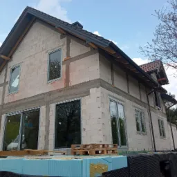 Surowy stan domu jednorodzinnego z zamontowanymi oknami z czarnymi ramami, widoczne ocieplenie fundamentów i palety drewniane.