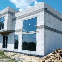 Surowy budynek z betonu komórkowego w trakcie budowy z zamontowanymi oknami z widocznymi refleksami nieba i drzew, na pierwszym planie stos desek i gruz.