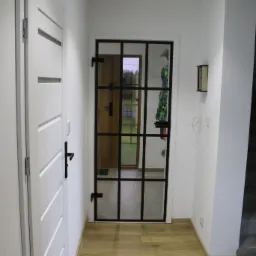 Wnętrze domu z dwiema białymi drzwiami, jedne z czarną ramą i szprosami, widok na zewnątrz, podłoga imitująca drewno, minimalistyczny styl.