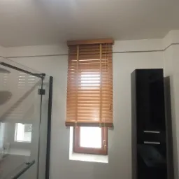 Jasne drewniane żaluzje poziome w oknie łazienki, widok z kabiny prysznicowej na tle białych płytek i czarnej szafki.