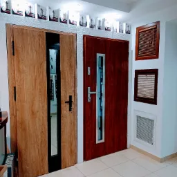 Ekspozycja dwóch różnych modeli drzwi zewnętrznych: jedne z jasnego drewna z czarnym wykończeniem i przeszkleniem, drugie z ciemnego drewna z wąskim, pionowym przeszkleniem i srebrnym okuciem,...