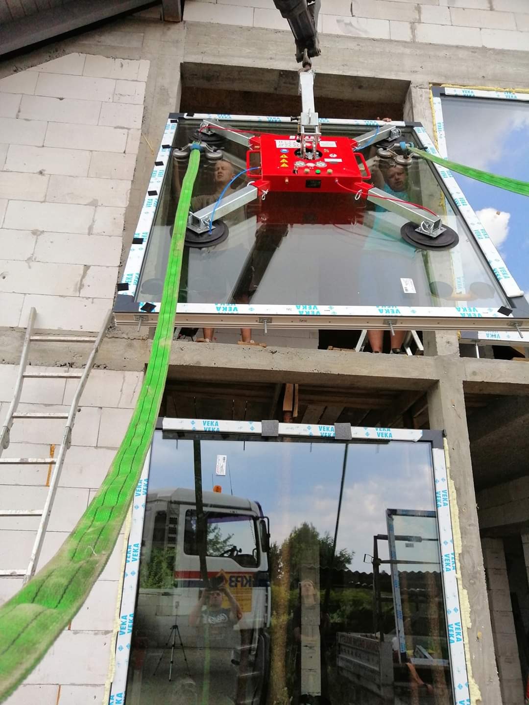 Montaż dużego okna z użyciem przyssawki próżniowej na ramie, widoczne liny zabezpieczające i odblask ciężarówki w szybie okna.