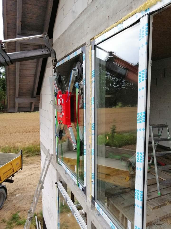 Montaż okien z profili z niebieską folią ochronną, przy użyciu specjalistycznego podnośnika próżniowego na budowie w Kraśniku. W tle widoczne pole i drabina.