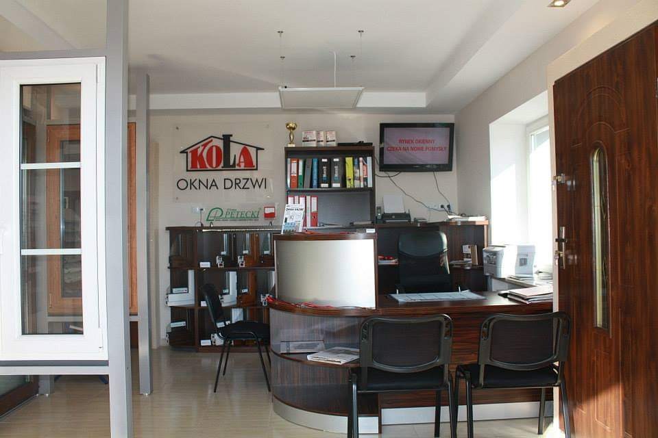 Wnętrze biura firmy oferującej stolarkę okienną i drzwiową, z ekspozycją okna z białymi ramami i drewnianej drzwi wejściowej po prawej stronie, za ladą recepcyjną widoczny regał z próbkami.