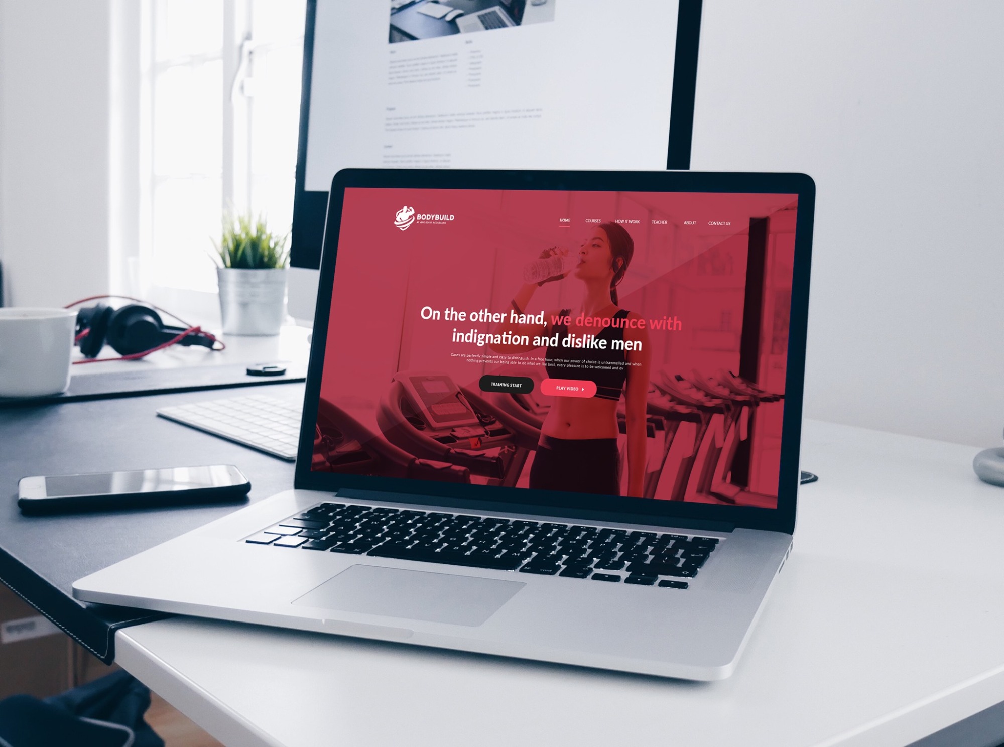 Laptop wyświetlający stronę internetową BODYBUILD z motywem fitness, na biurku obok telefonu i kubka, drugi monitor w tle.