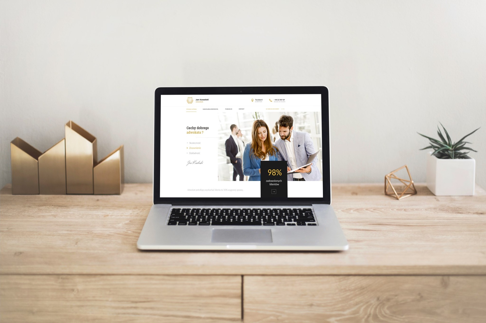 Laptop na drewnianym biurku prezentuje stronę internetową kancelarii adwokackiej z informacjami o skuteczności i zadowoleniu klientów, obok złota dekoracja i roślina w białej doniczce.