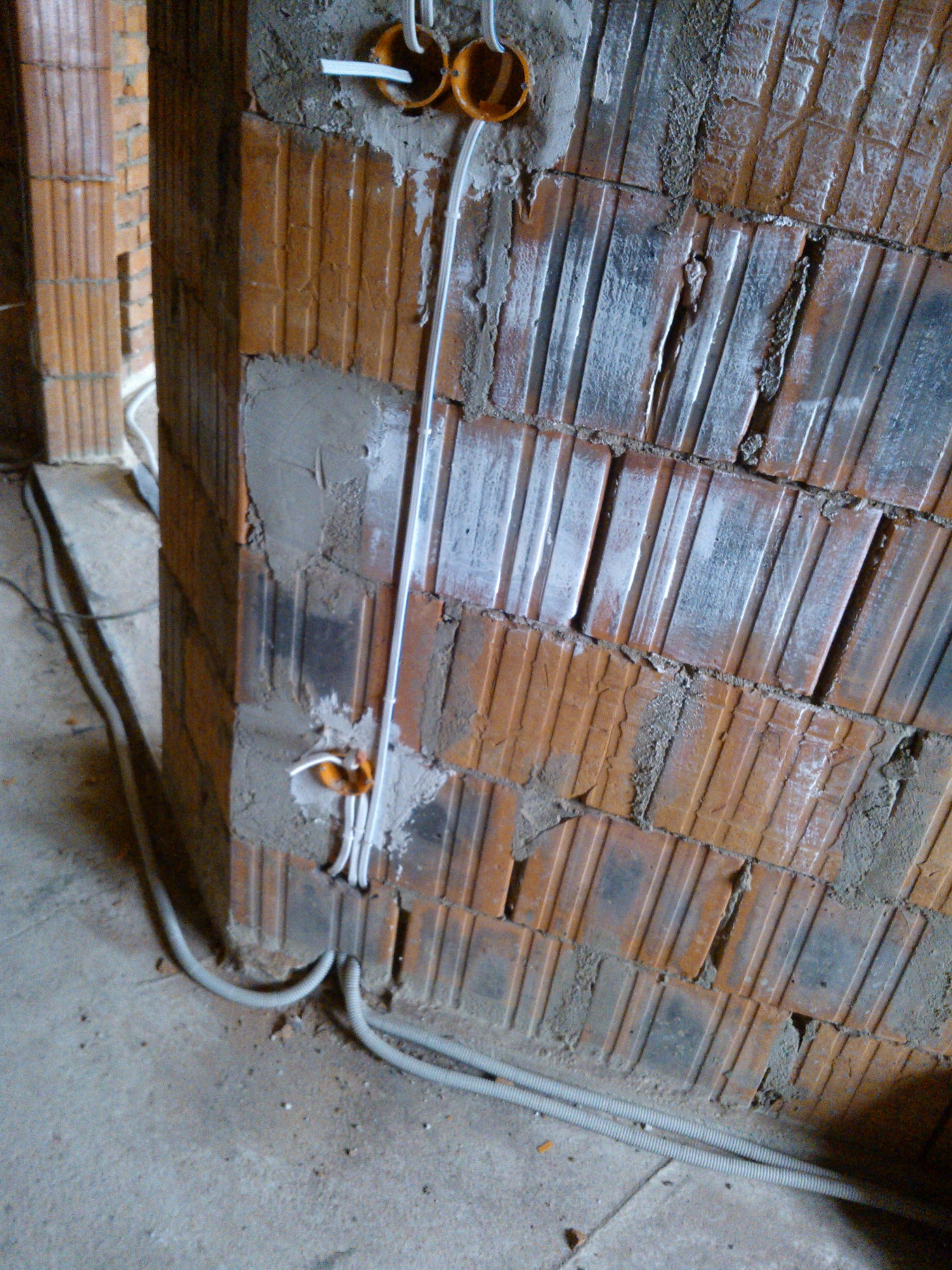 Instalacja przewodów elektrycznych w ścianie z cegły ceramicznej, widoczne puszki instalacyjne i peszle przy podłodze.