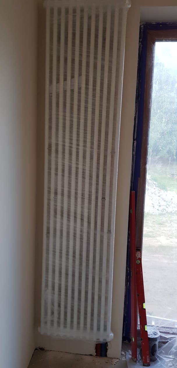 Nowy, biały grzejnik żeberkowy, zabezpieczony folią ochronną, stoi obok okna z widoczną zielenią na zewnątrz; obok grzejnika oparta poziomica.