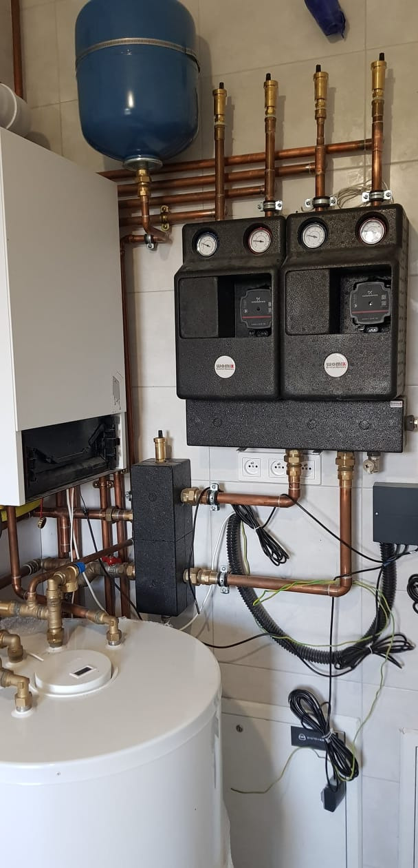 Złożony system instalacji grzewczej z kotłem, zbiornikiem, pompami obiegowymi i licznymi rurami miedzianymi, zamontowany na tle kafelkowanej ściany.