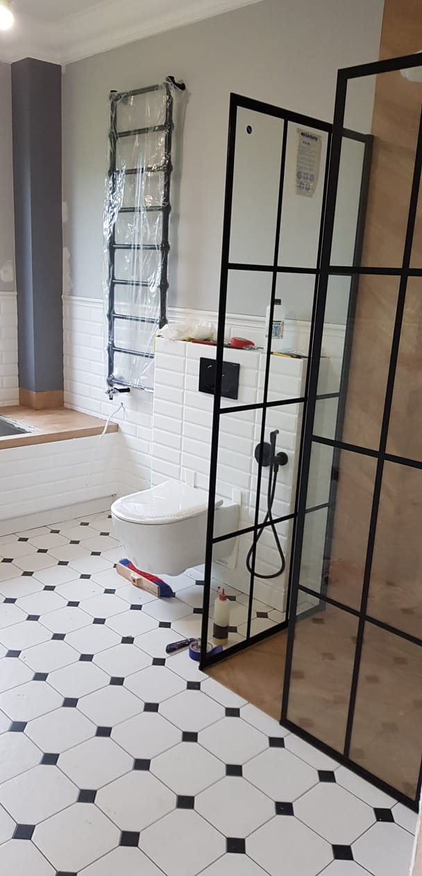 Montaż kabiny prysznicowej typu loft w łazience z białą armaturą i czarno-białą podłogą w karo, widoczny grzejnik drabinkowy w folii ochronnej.