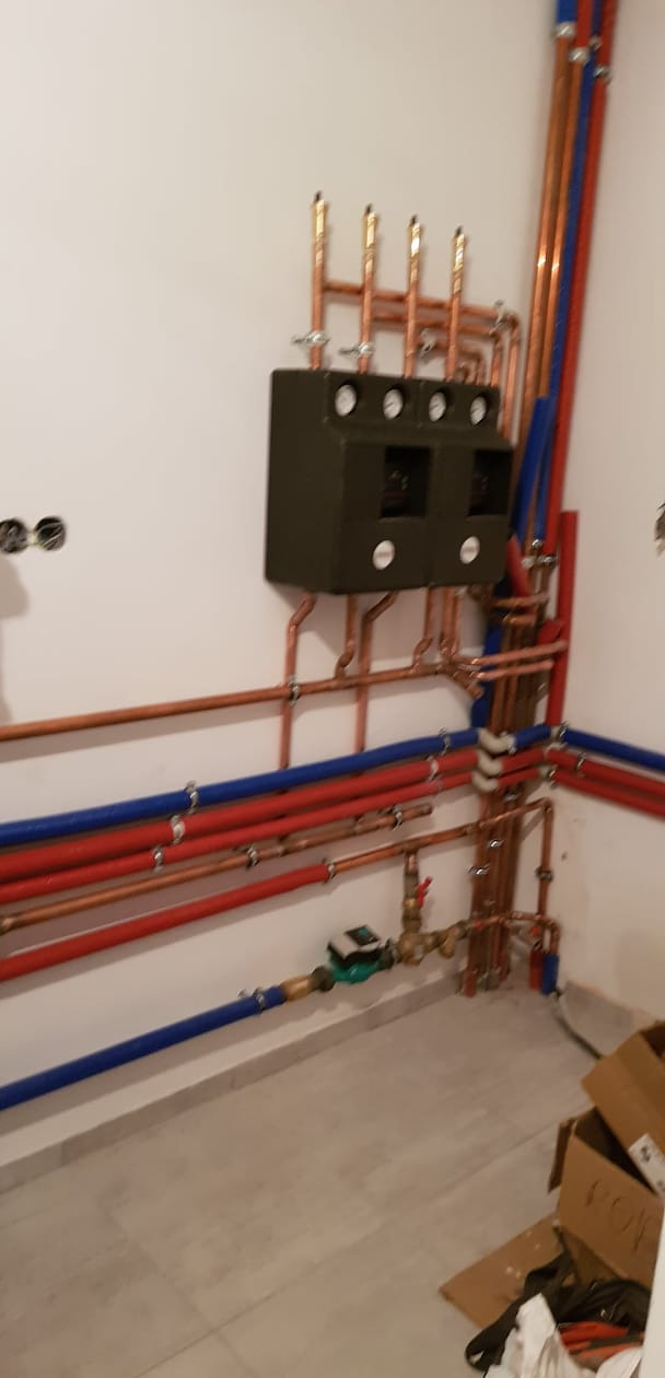 Rozdzielacz centralnego ogrzewania z licznymi rurami miedzianymi i plastikowymi w kolorach czerwonym i niebieskim, zamontowany na białej ścianie w nowym budynku, widoczne niedokończone prace...