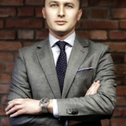 Portret pewnego radcy prawnego w szarym garniturze na tle ceglanej ściany, z zegarkiem na nadgarstku i złożonymi rękami.