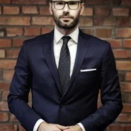 Portret mężczyzny w okularach i granatowym garniturze na tle ściany z czerwonej cegły, ręce złożone przed sobą.