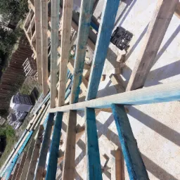 Widok z góry na konstrukcję drewnianą z belek pomalowanych na niebiesko, prawdopodobnie w trakcie budowy, z widocznymi narzędziami i elementami konstrukcyjnymi na jasnym tle. W tle widoczny ogród...