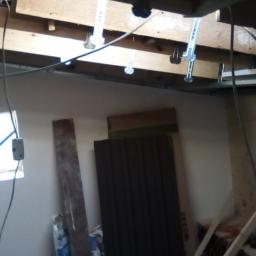 Pew-bud - Poddasze w trakcie remontu: widoczne metalowe profile sufitowe, częściowo otynkowane ściany, okno z niebieską taśmą zabezpieczającą, materiały budowlane w tle.