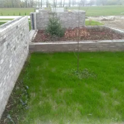 Murowana ściana z szarej cegły klinkierowej, tworząca obramowanie rabaty z iglakami i krzewami na trawniku, widok z perspektywy niskiego kąta.