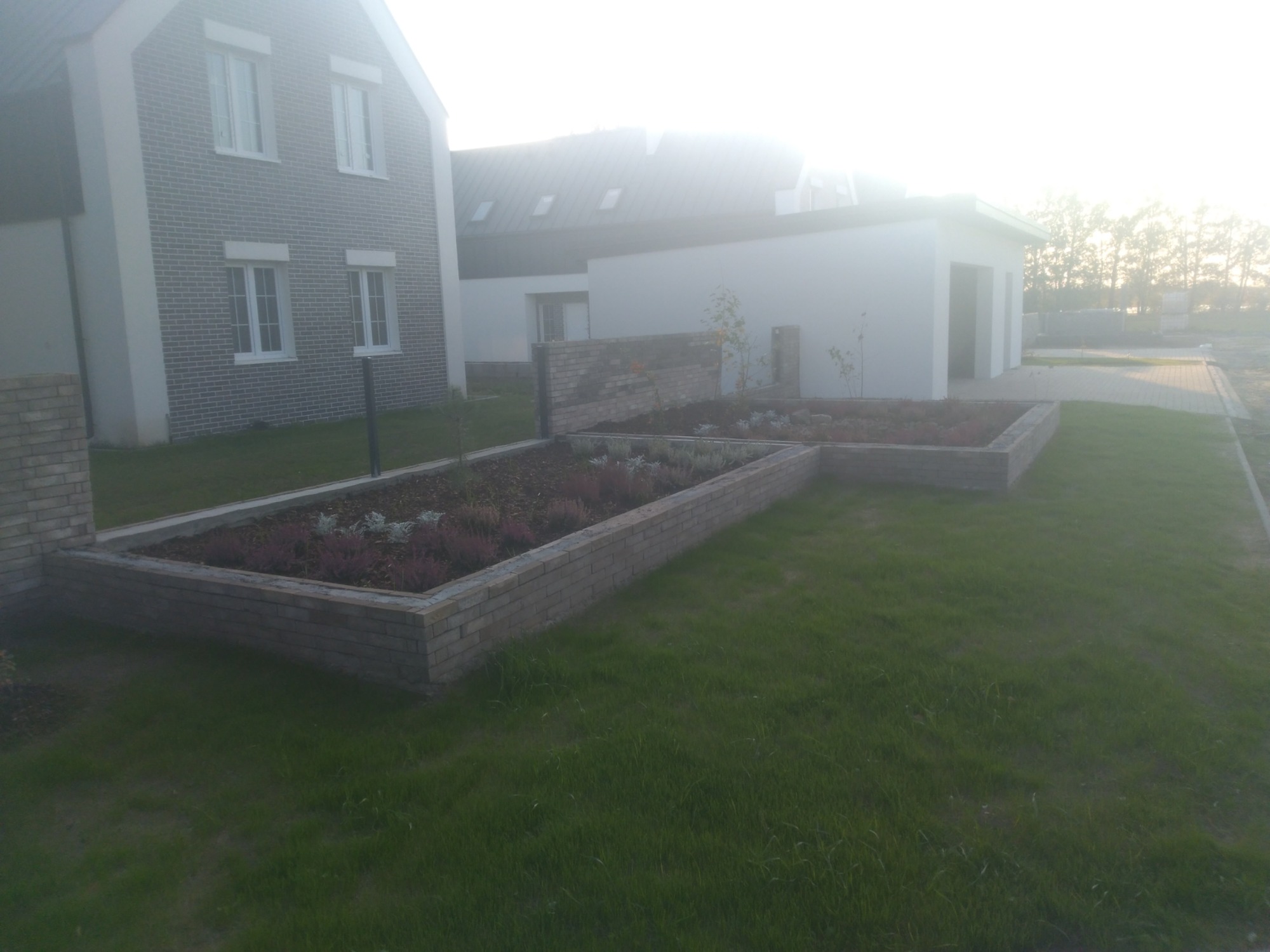 Dwa podwyższone kwietniki z cegły, obsadzone wrzosami i roślinami okrywowymi, na tle nowoczesnego domu z szarej cegły i białego garażu, w porannym świetle.