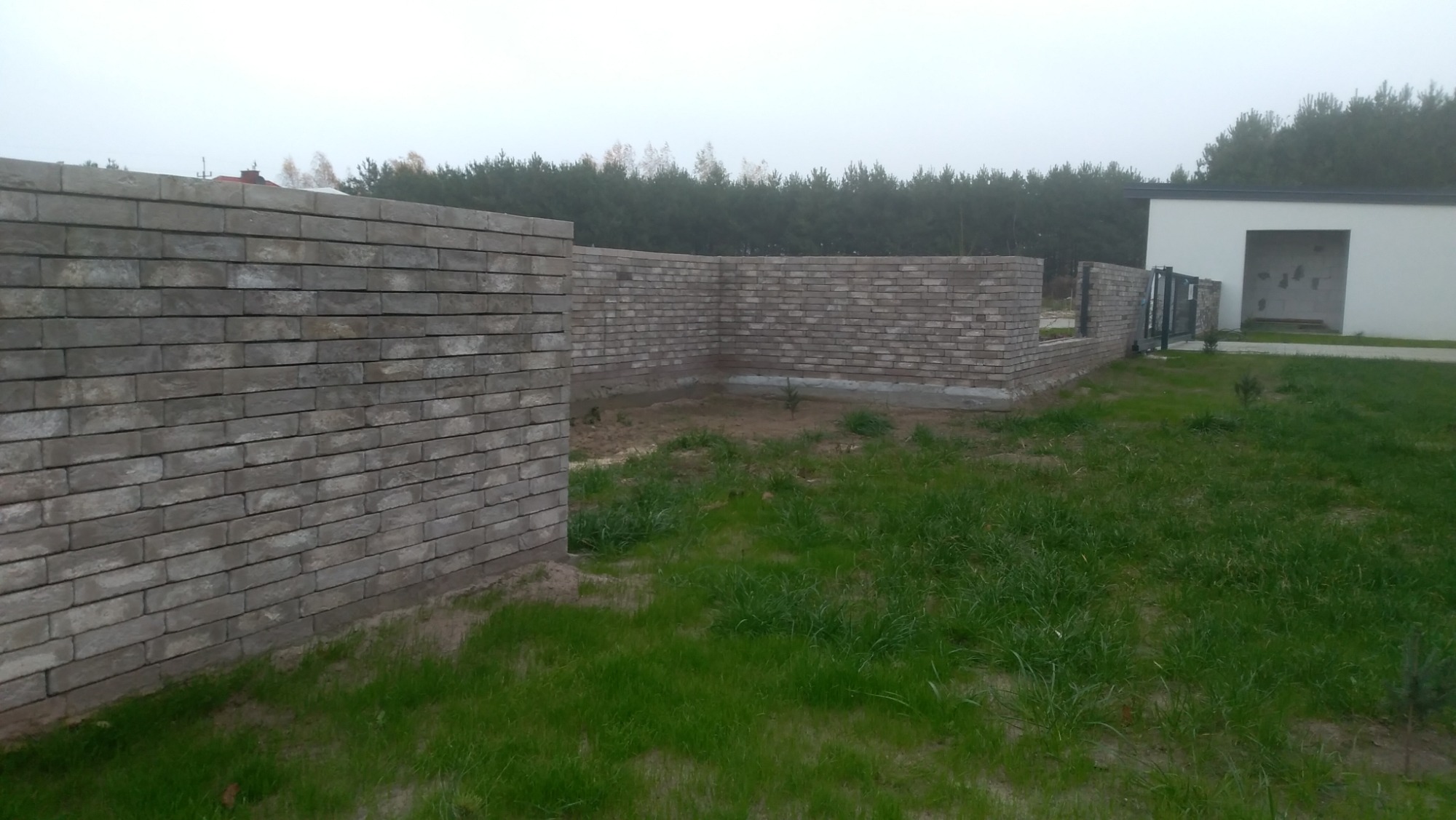 Wznoszone mury z szarej cegły klinkierowej na działce, widoczny fragment budynku w tle i brama wjazdowa.
