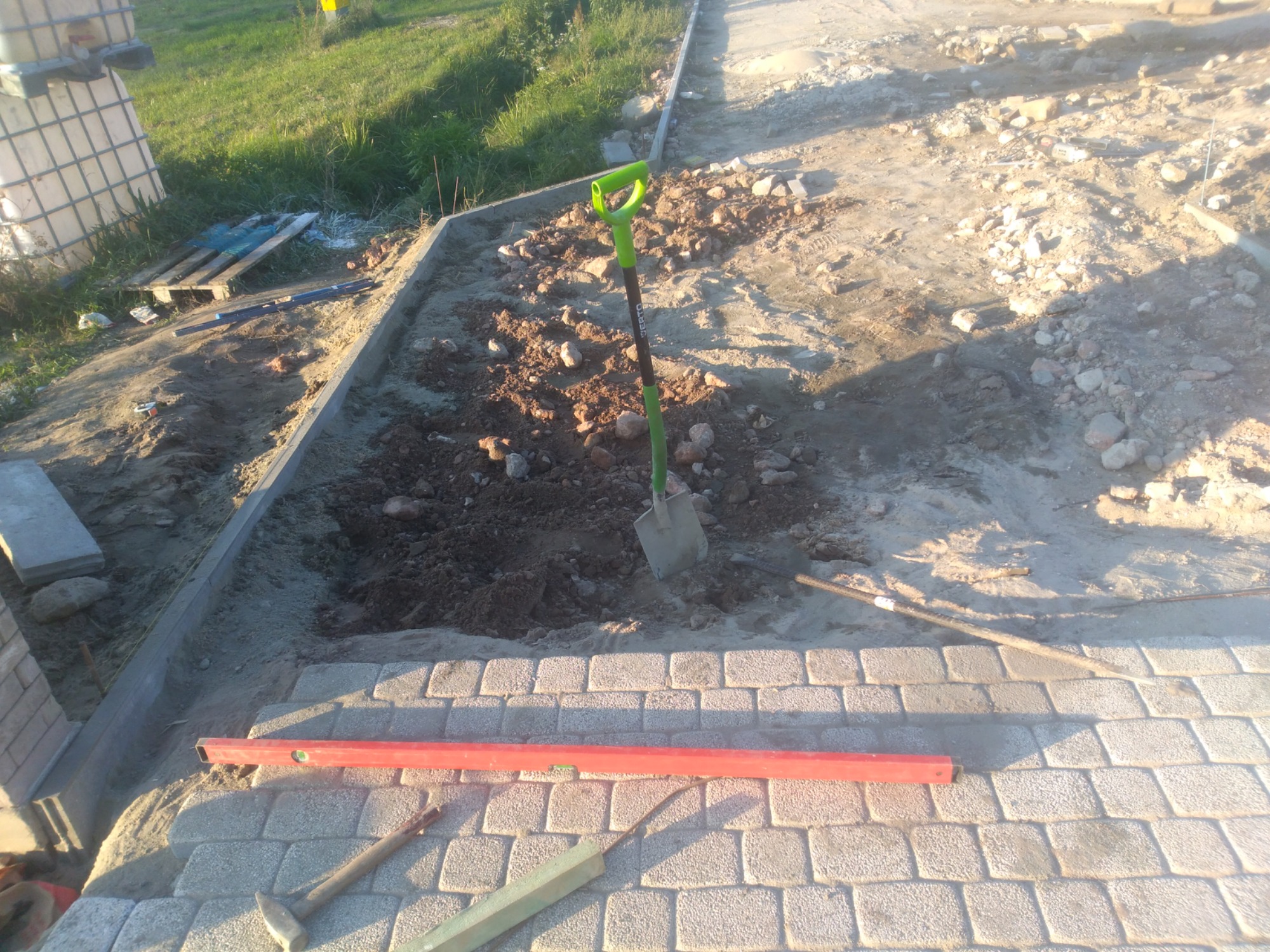 Układanie kostki brukowej na podsypce piaskowej, widoczna poziomica, łopata z zieloną rączką i młotek brukarski