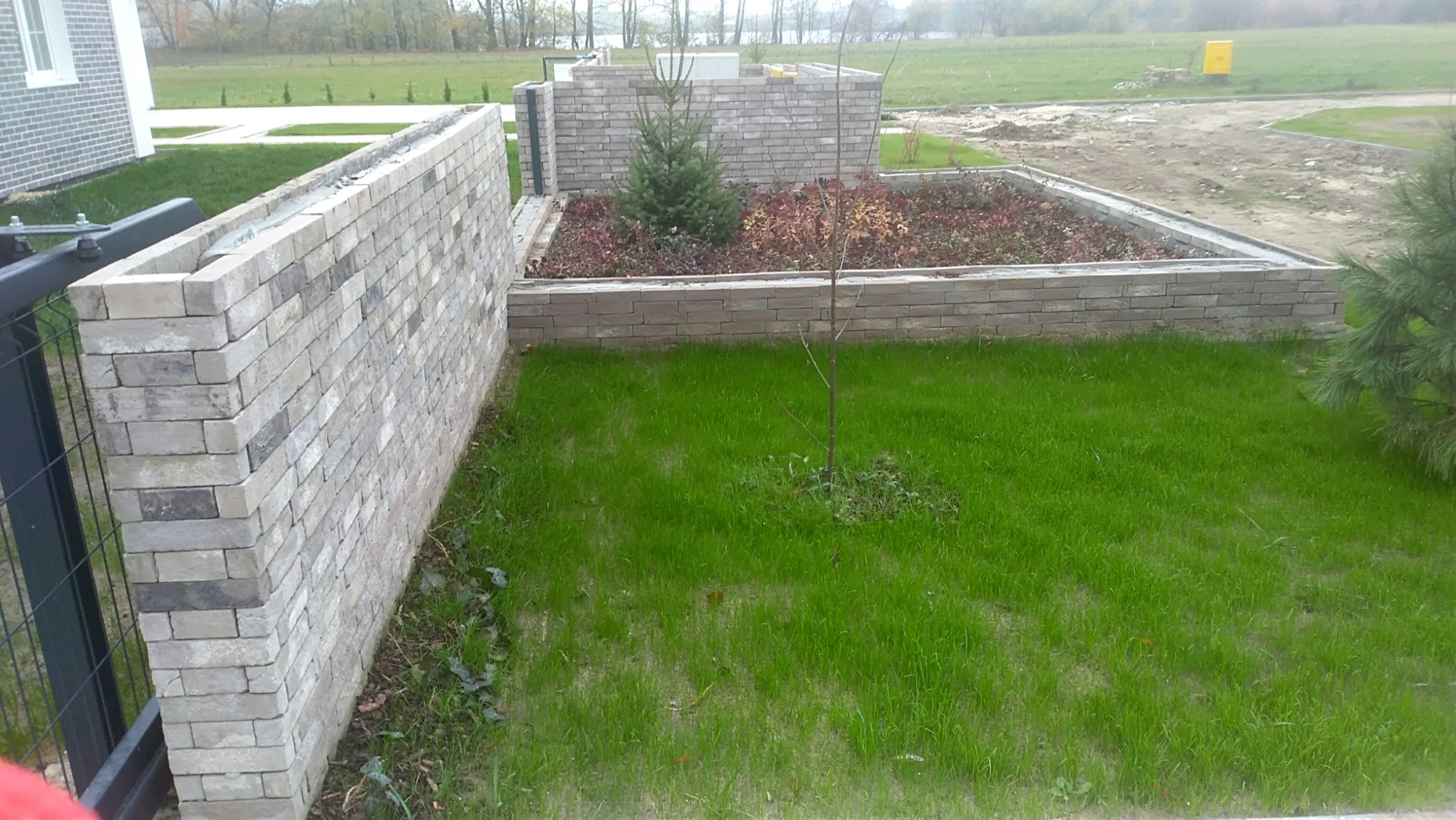 Murowana ściana z szarej cegły klinkierowej, tworząca obramowanie rabaty z iglakami i krzewami na trawniku, widok z perspektywy niskiego kąta.