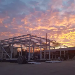 Konstrukcja stalowa hali w trakcie montażu, na tle dramatycznego, pomarańczowo-różowego zachodu słońca. Widoczne elementy konstrukcyjne, zwoje kabli oraz sąsiedni budynek.