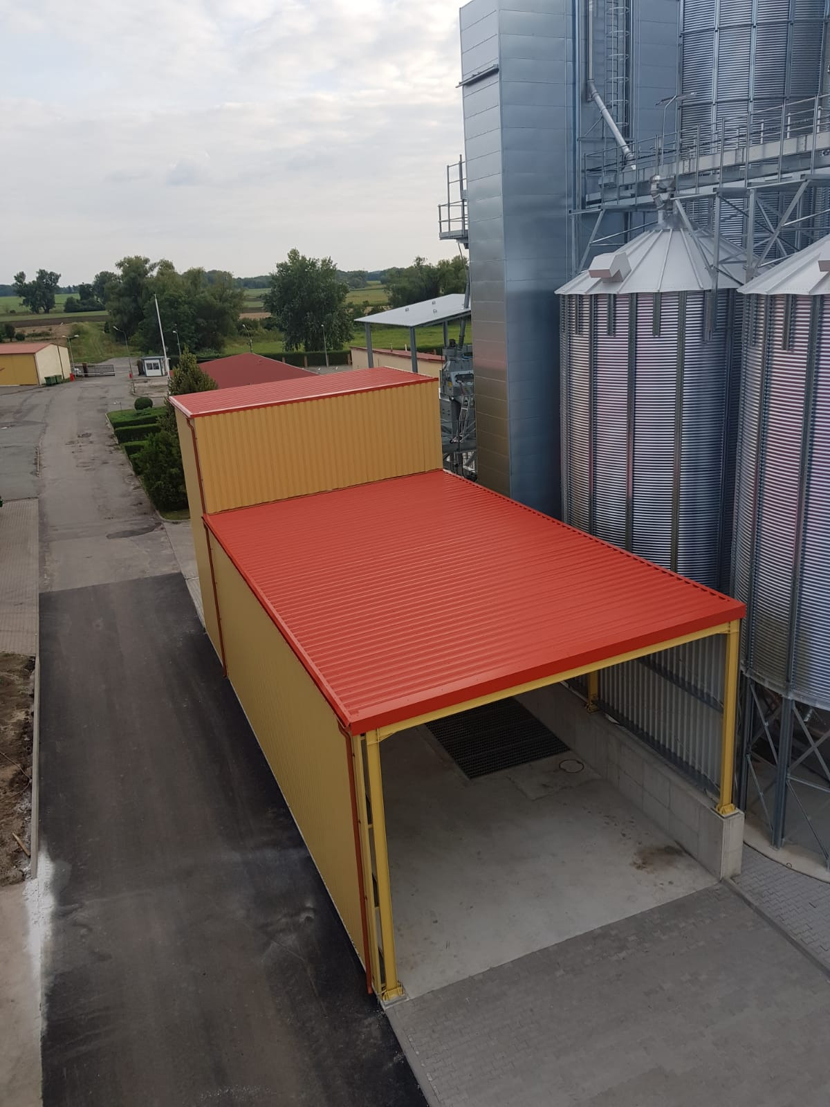 Widok z góry na halę magazynową z czerwonym dachem i żółtymi ścianami z blachy trapezowej, przylegającą do silosu zbożowego z metalu falistego, na tle krajobrazu wiejskiego.