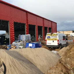 Przygotowania do wylewki posadzki: sterta piasku, worki z zaprawą i agregat w tle czerwonej hali magazynowej, obok zaparkowany bus firmy budowlanej.