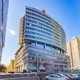 Nowoczesny budynek biurowy z zaokrągloną fasadą i logo firmy na dachu, widok z dołu na tle błękitnego nieba, zaparkowane samochody na pierwszym planie.