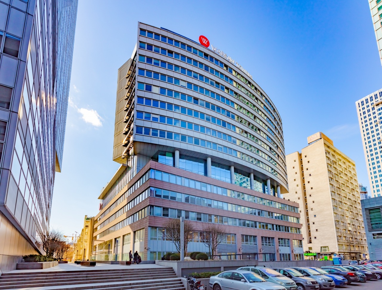 Nowoczesny budynek biurowy z zaokrągloną fasadą i logo firmy na dachu, widok z dołu na tle błękitnego nieba, zaparkowane samochody na pierwszym planie.