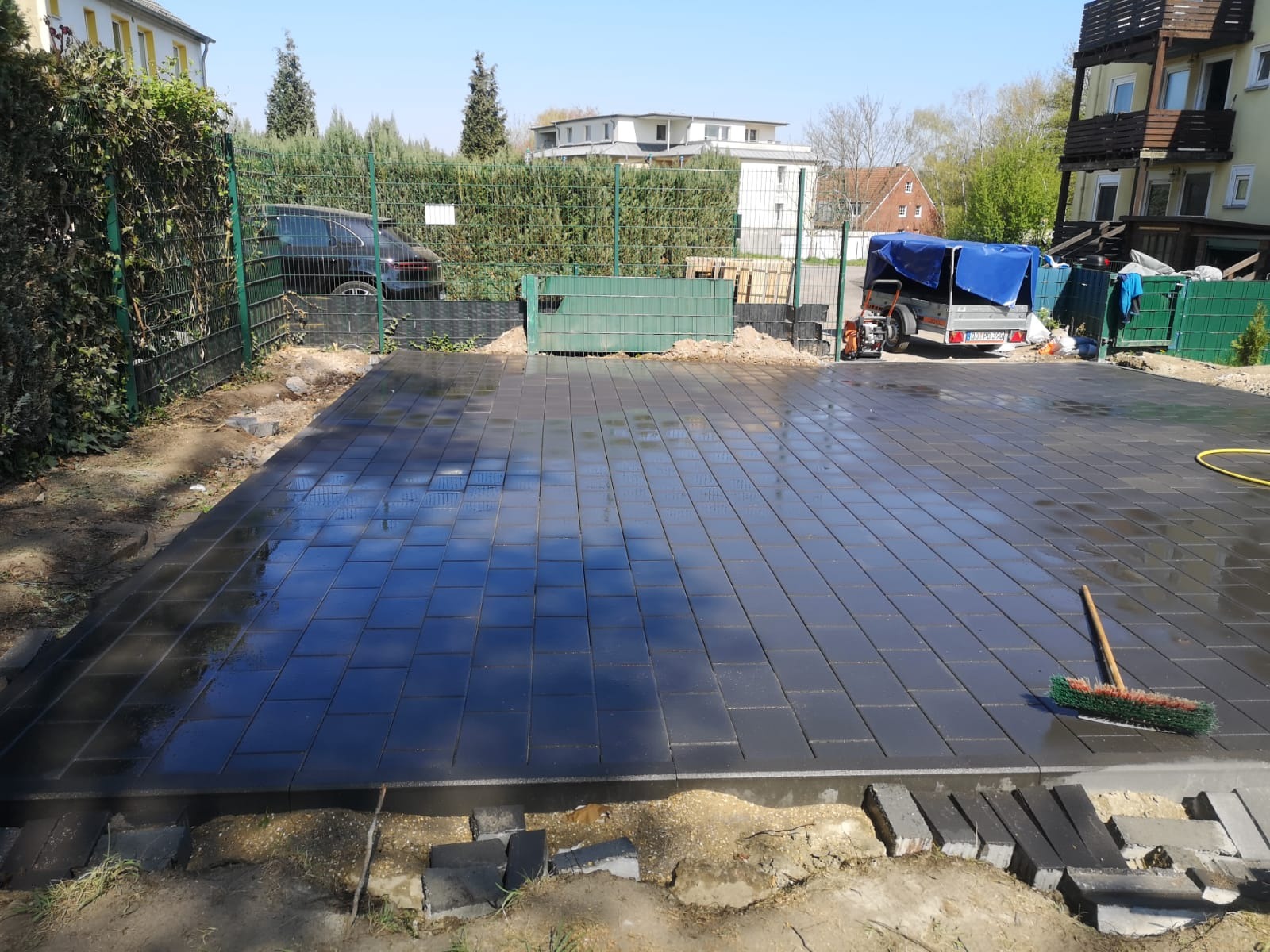 Świeżo ułożona, mokra, grafitowa kostka brukowa na posesji z widocznymi elementami przygotowania podłoża, szczotką do zamiatania i przyczepką w tle.