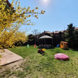 Słoneczny plac zabaw na trawie z żółtą forsycją w rogu, widoczna altana, domek, zjeżdżalnie i zabawki