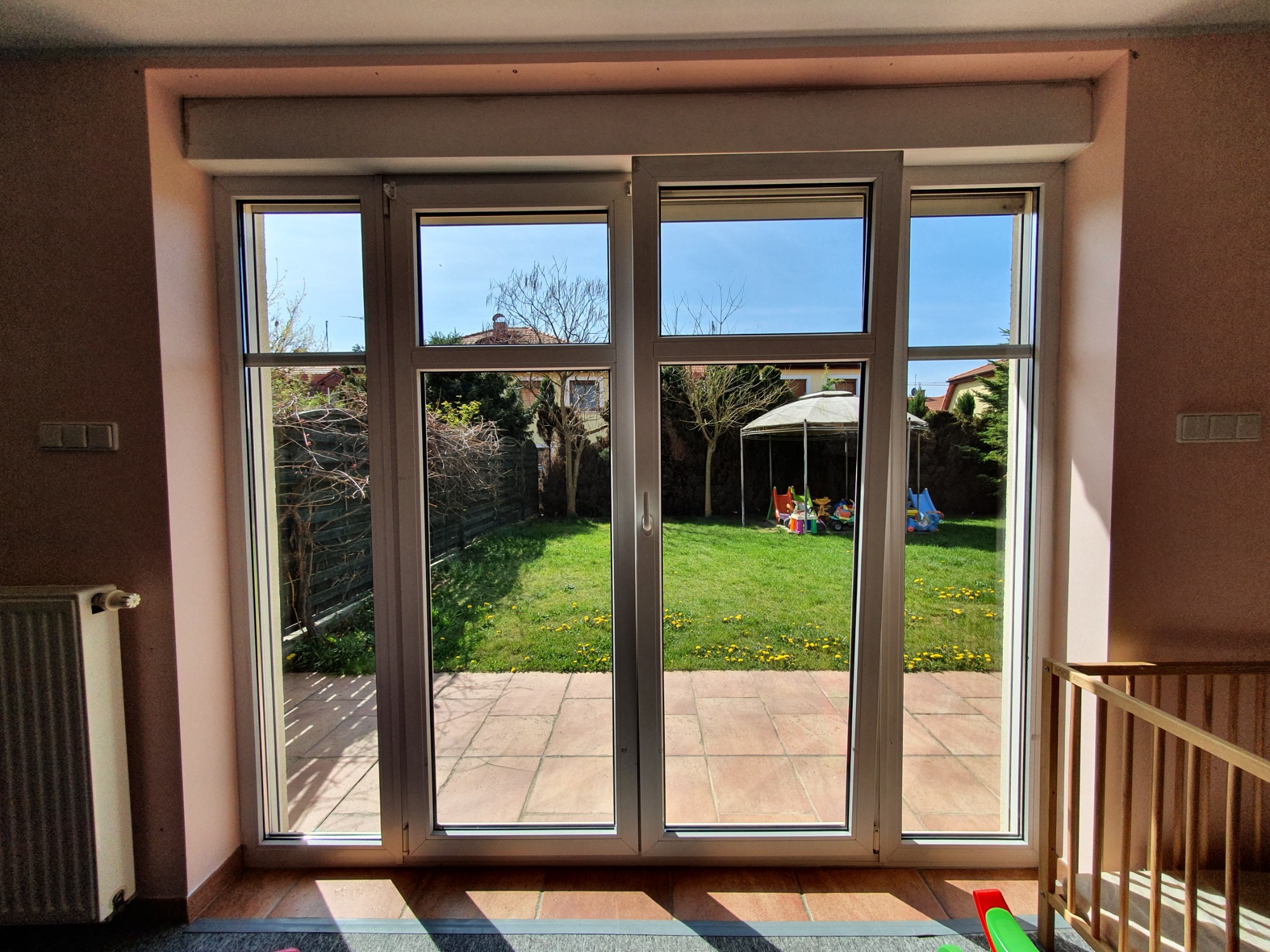 Widok z pokoju dziecięcego przez szerokie, przesuwne okno na zielony ogród z placem zabaw i altaną