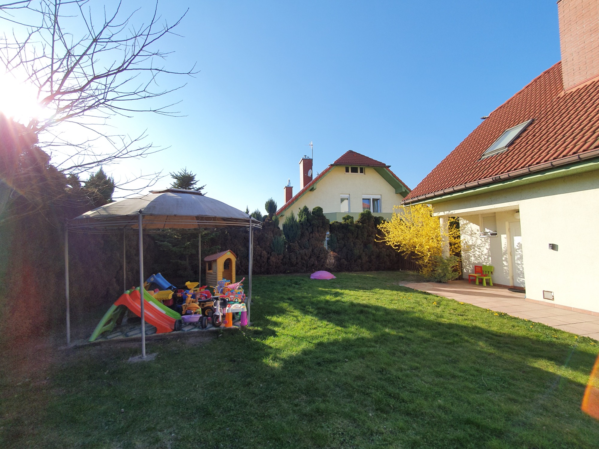 Słoneczny ogród z metalowym zadaszeniem nad plastikową zjeżdżalnią i zabawkami, trawnik, dom jednorodzinny w tle.