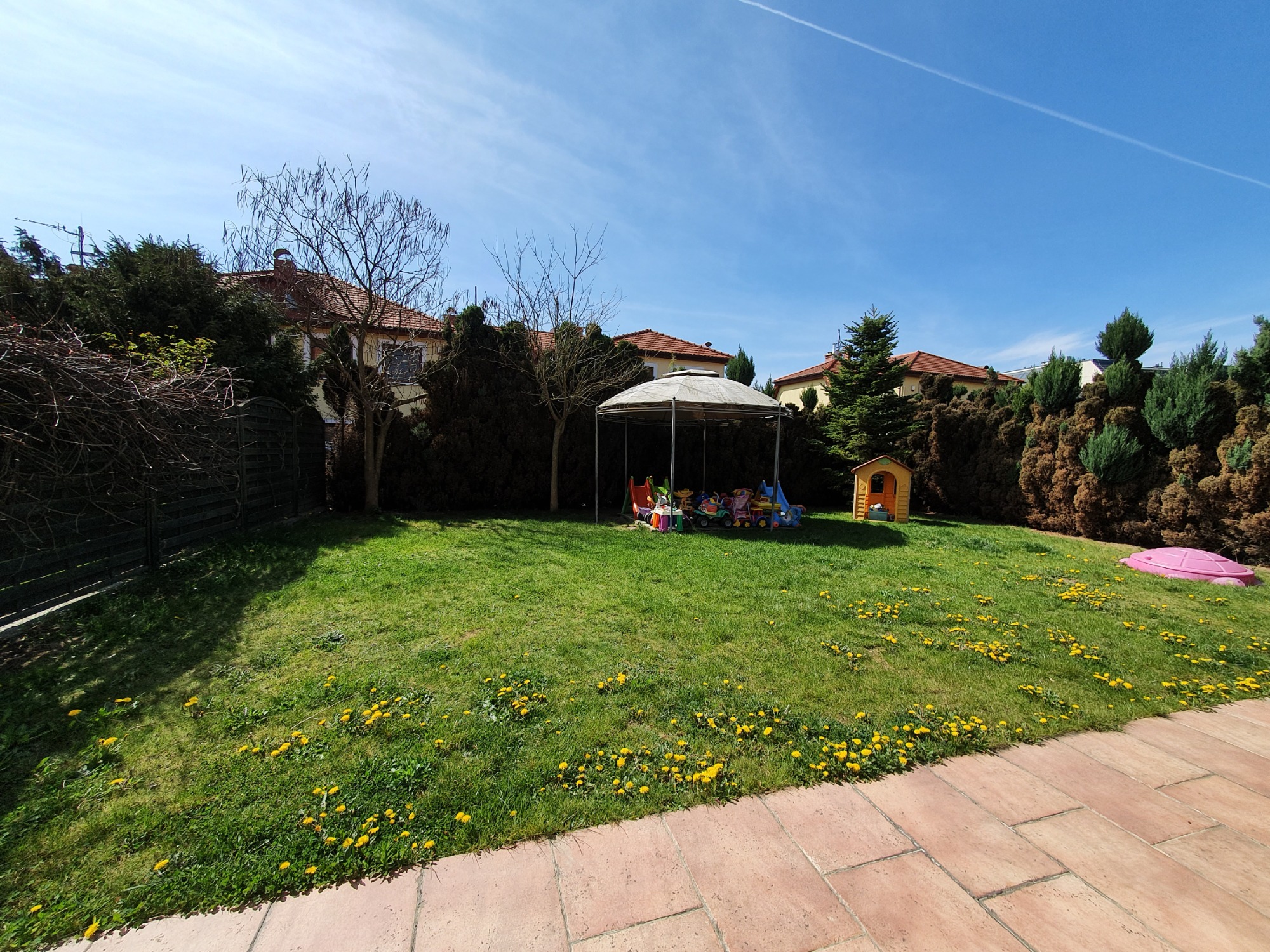 Przestronny, zielony ogród z placem zabaw pod zadaszeniem, małym domkiem i piaskownicą w słoneczny dzień.