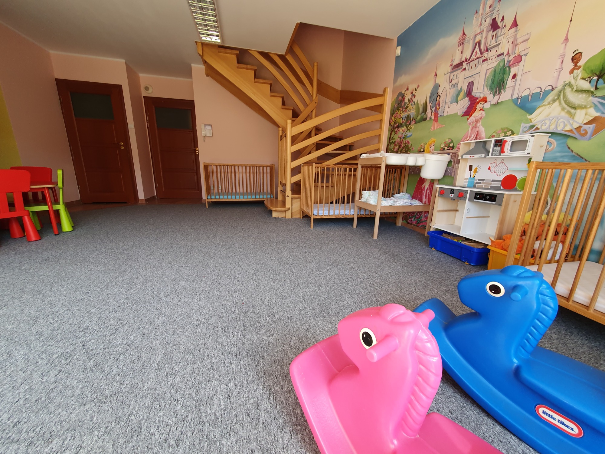Wnętrze żłobka z drewnianymi schodami, łóżeczkami, zabawkami i ścianą z motywem zamku księżniczek, z przodu widoczne dwa plastikowe koniki na biegunach: różowy i niebieski.