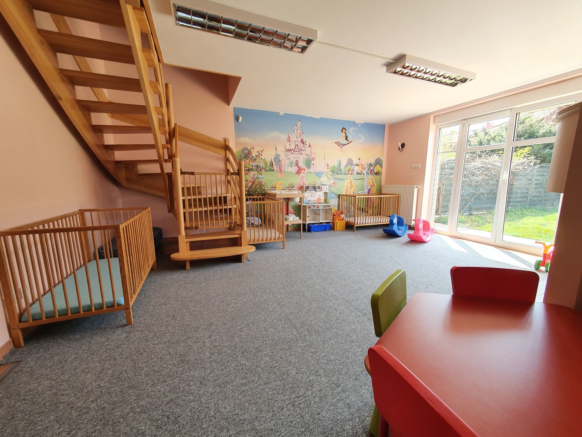 Jasny pokój z drewnianymi łóżeczkami dziecięcymi, drewnianymi schodami, stolikiem z krzesłami oraz ścianą z bajkowym motywem zamku i postaci.