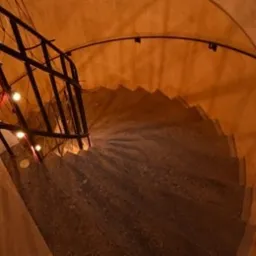 Wewnętrzna, spiralna klatka schodowa z betonowymi stopniami, oświetlona ciepłym światłem, z metalową balustradą, widok z góry.