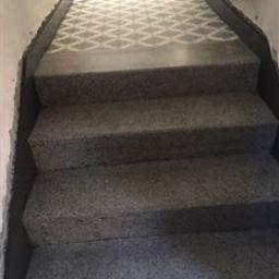 Polfloormax - Betonowe schody z szarymi stopniami i wzorzystą posadzką na podeście, widok z dołu w kierunku jasnego wyjścia.