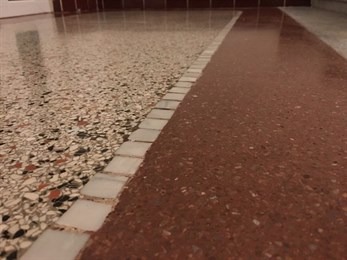 Polerowana posadzka lastryko z wyraźnym podziałem na dwie sekcje kolorystyczne: beżowo-szara i czerwona, rozdzielone białym pasem z kwadratowych płytek. Powierzchnia odbija światło, ukazując drobne...