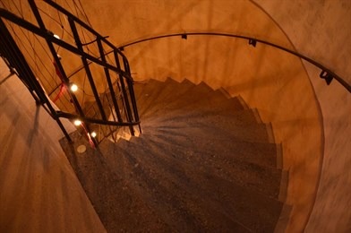 Wewnętrzna, spiralna klatka schodowa z betonowymi stopniami, oświetlona ciepłym światłem, z metalową balustradą, widok z góry.
